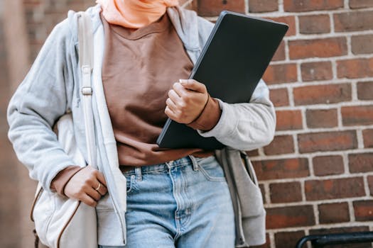 Unrecognizable woman in casual outfit holding laptop against brick wall outdoors.